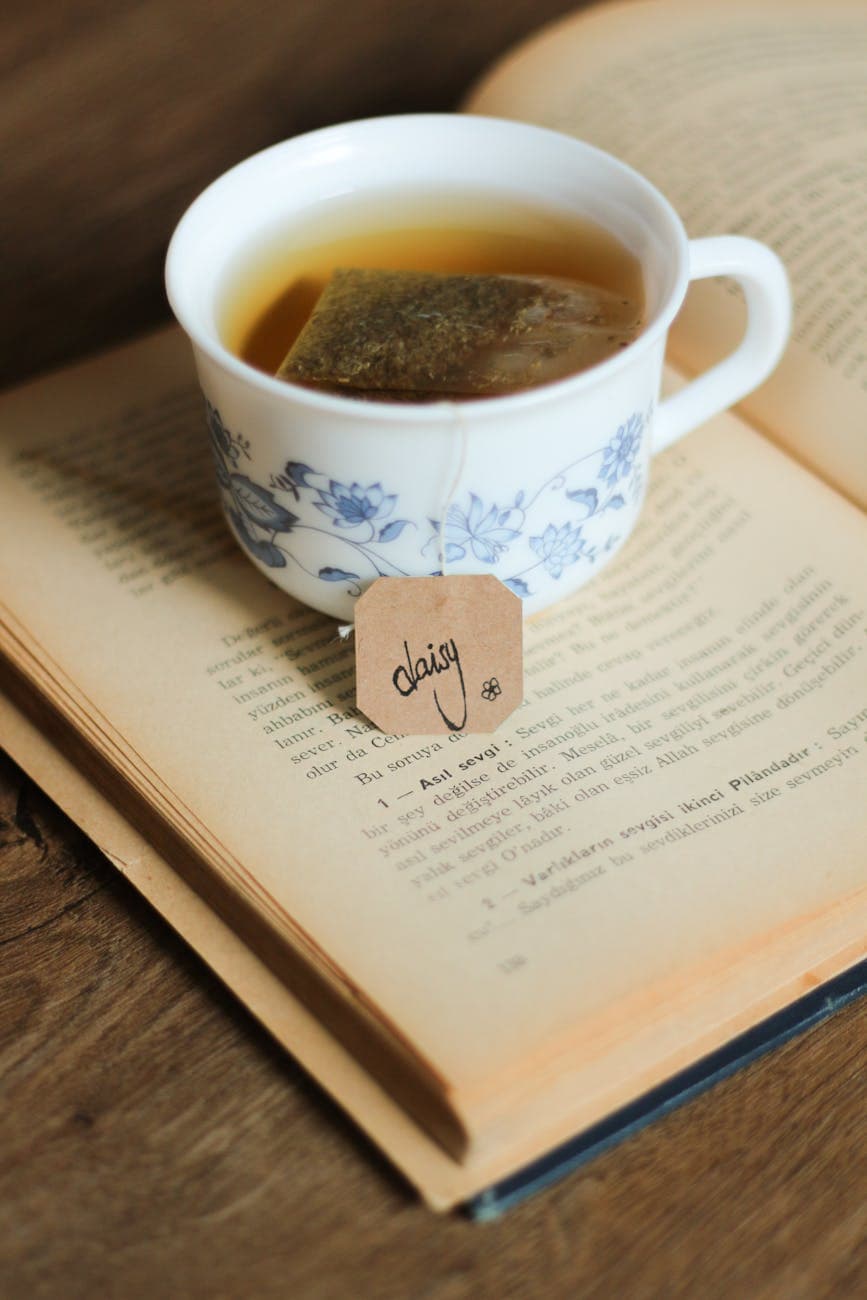 A cup of tea with a tea bag steeping beside an open book, with a string and label visible, representing the classic non-heat-seal European tea bag format