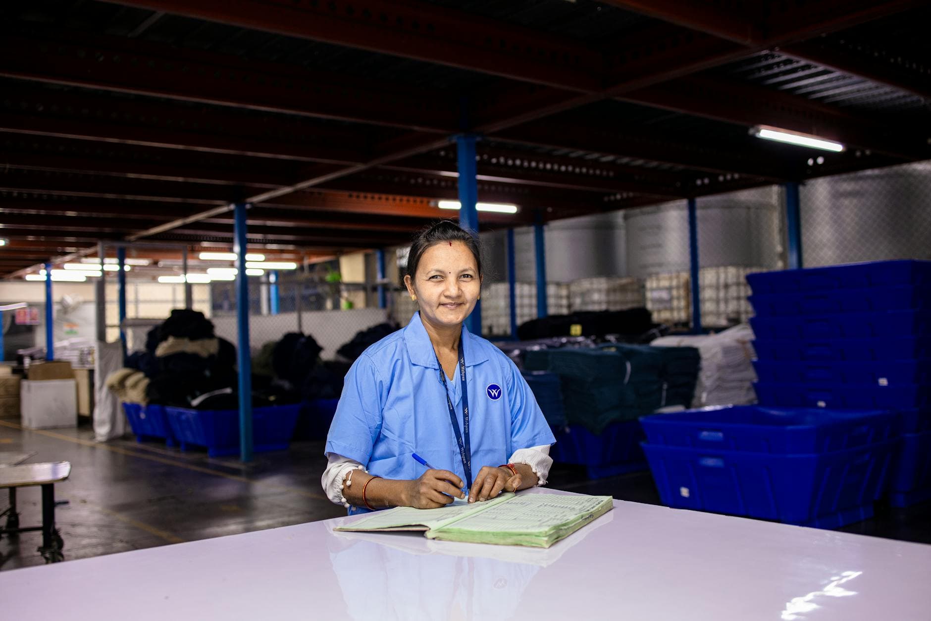 Female worker in uniform writing in a notebook inside a warehouse, representing quality control and production documentation
