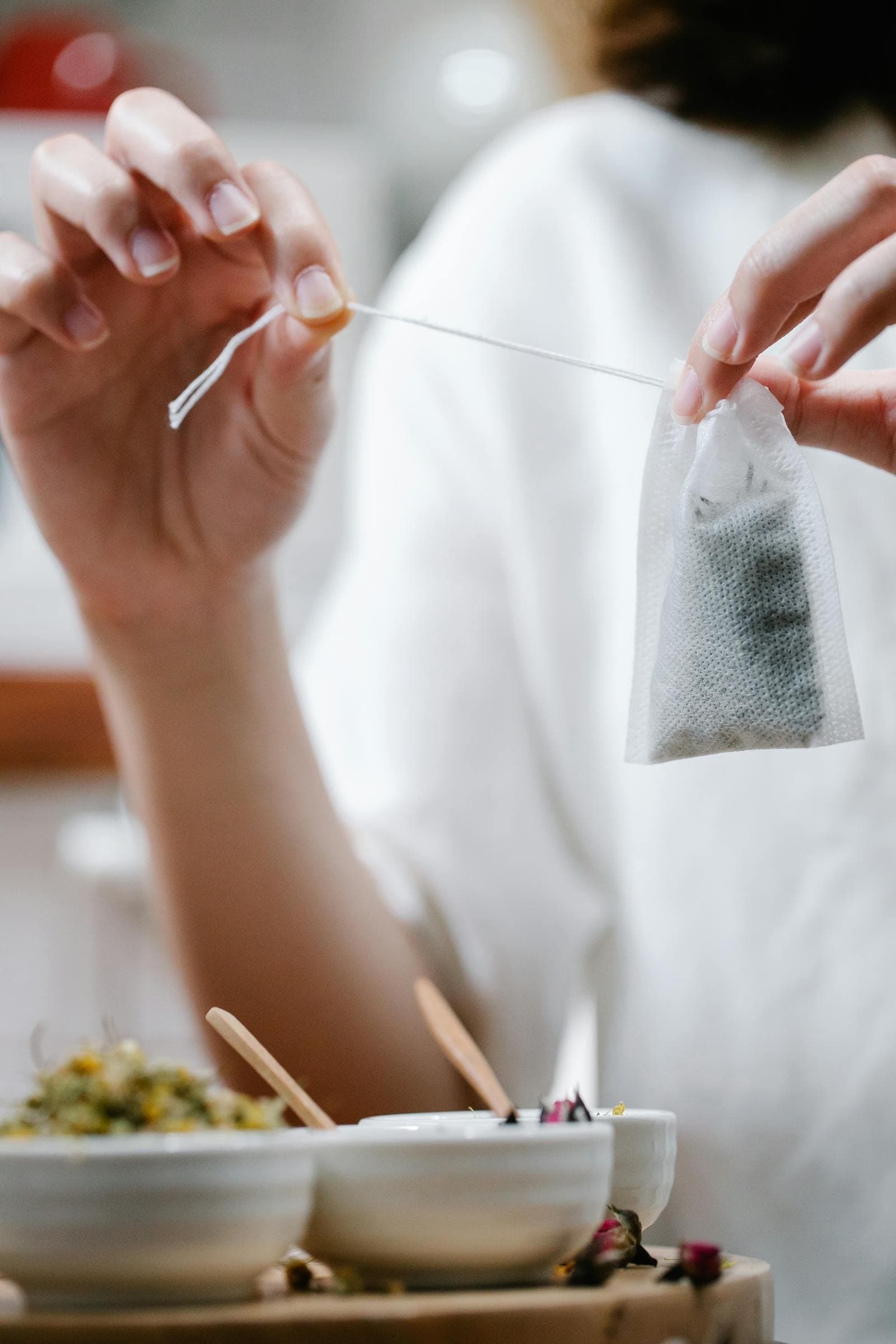 A hand lifting a single-string paper tea bag above a tray of dried herbs and rose petals